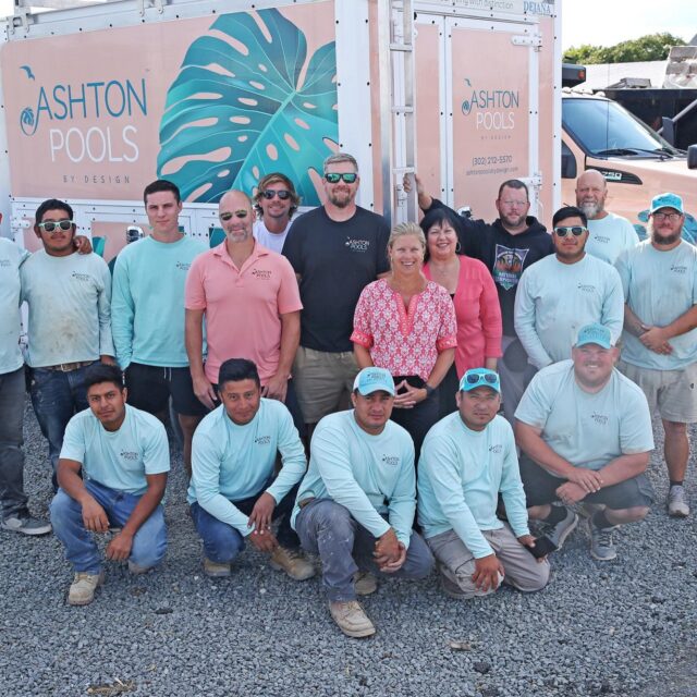 A group of 18 people pose in front of two Ashton Pools by Design trucks in a gravel lot. Most are wearing matching light blue shirts with the company logo, ready to create luxury pools and custom spas for exceptional outdoor living.