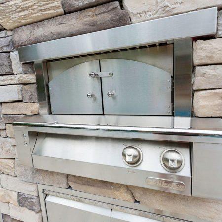 Stainless steel outdoor pizza oven with two control knobs, built into a stone wall—perfect for outdoor kitchens near luxury pools or custom spas.