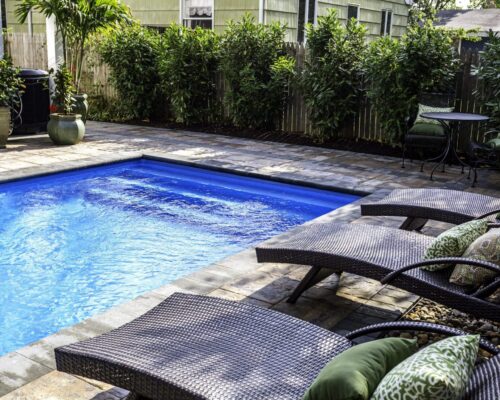Rectangular backyard pool with lounge chairs, potted plants, and a small table with chairs, surrounded by privacy hedges and a stone patio—perfect for enjoying the luxury pools lifestyle or adding custom spas to your outdoor retreat.