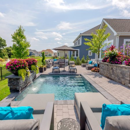 Modern backyard with a small fiberglass pool, gray patio furniture, flower arrangements, stonework, and a gray house in the background—perfect for outdoor living under a partly cloudy sky.