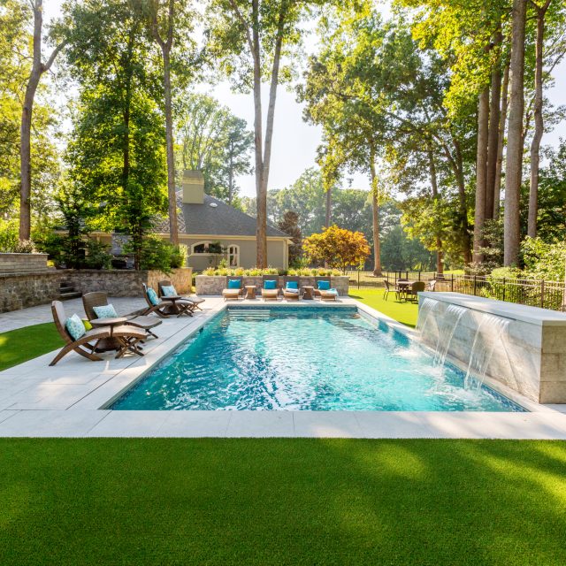 Backyard with a rectangular fiberglass pool, waterfall feature, lounge chairs, and artificial grass, surrounded by trees and a large house.