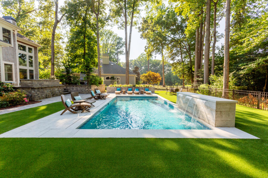 Backyard with a rectangular fiberglass pool, waterfall feature, lounge chairs, and artificial grass, surrounded by trees and a large house.