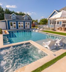 A backyard oasis featuring luxury pools, a hot tub, lounge chairs, outdoor dining with umbrellas, and a blue house with a guest house in the background—perfect for elevated outdoor living.