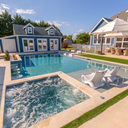 A backyard oasis featuring luxury pools, a hot tub, lounge chairs, outdoor dining with umbrellas, and a blue house with a guest house in the background—perfect for elevated outdoor living.