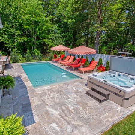 A backyard patio with a swimming pool, red lounge chairs under umbrellas, a hot tub, and surrounding greenery creates the perfect outdoor living space for relaxing or entertaining.