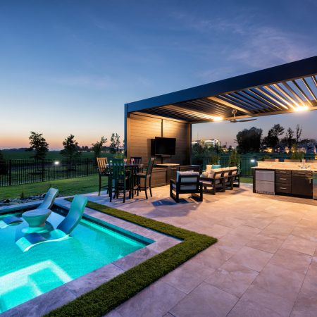 Modern backyard with illuminated fiberglass pool, lounge chairs in the water, outdoor seating area under a pergola, and built-in outdoor kitchen at dusk.