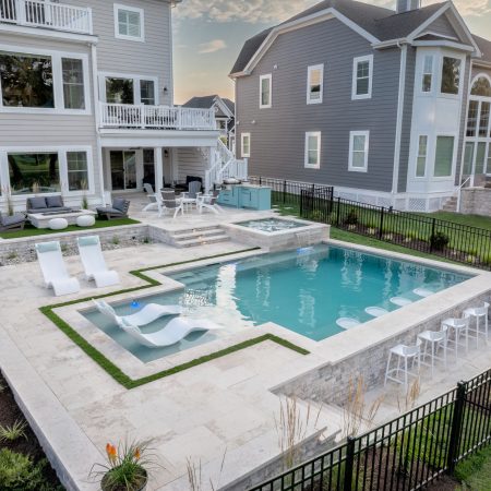 Modern backyard with a luxury pool, lounge chairs in the water, a hot tub, stylish outdoor living spaces, and two large houses in the background.