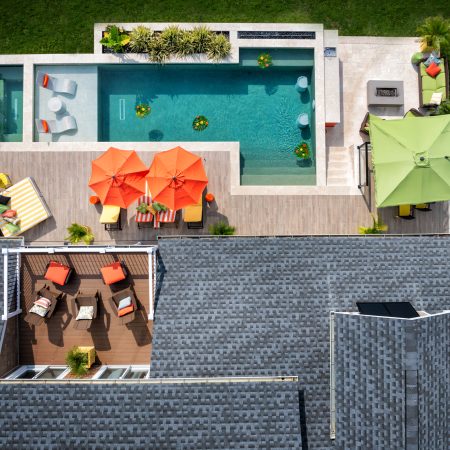 Aerial view of a backyard with a luxury inground pool, lounge chairs, orange and green umbrellas, wooden deck, and surrounding lawn.