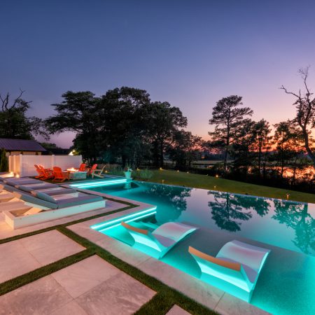 Modern backyard with illuminated infinity inground pool, sun loungers partially submerged in water, a lounge seating area, and a sunset view with trees and a reflecting pond in the background.